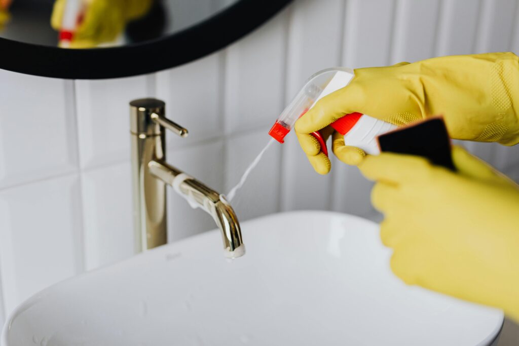 pexels-photo-4239130-4239130 From above anonymous housekeeper in yellow rubber gloves spraying cleaner to chrome water faucet in bathroom