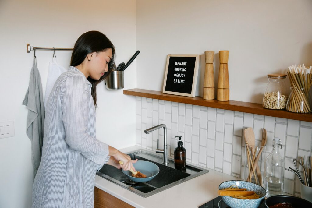 pexels-photo-7262358-7262358 Side view of Asian female with brush washing dishes at sink while doing housework in light kitchen with supplies at home